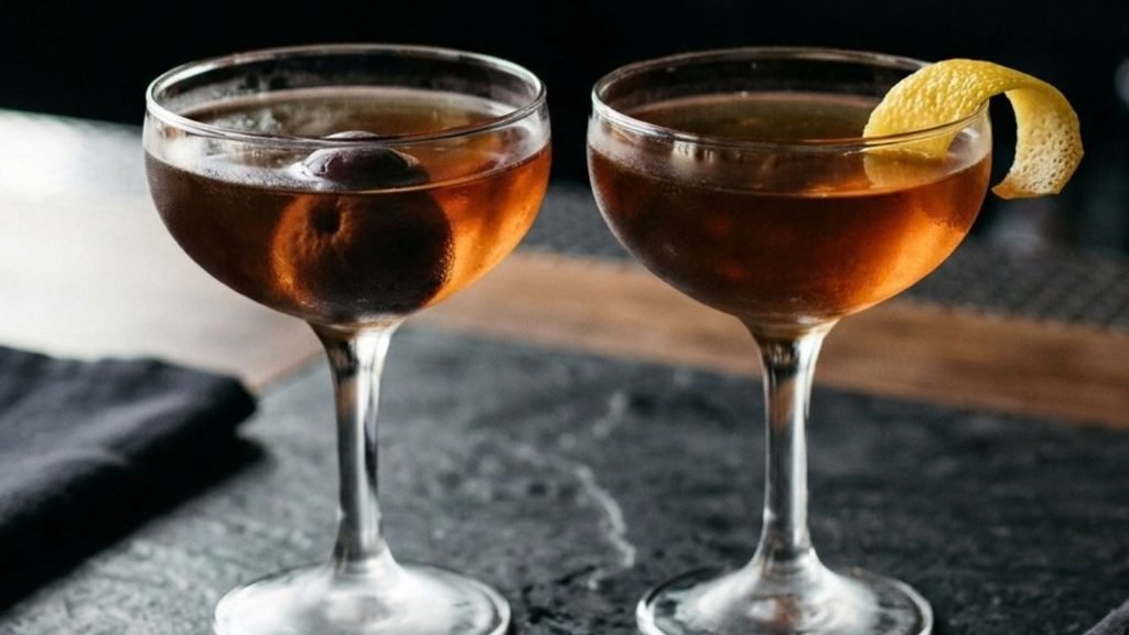 Two Manhattan cocktails, one garnished with a cherry and the other with a lemon peel.

